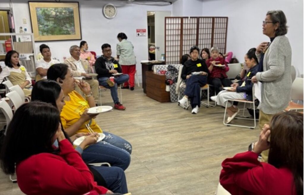 A group of approximately fifteen individuals are gathered in a brightly lit indoor space, seemingly engaged in a meeting or workshop. Most of the attendees are seated in chairs arranged in a semi-circular fashion, facing a person who appears to be speaking while standing. This individual is positioned towards the right side of the frame, holding papers in their hands. The attendees are diverse in appearance and are dressed in casual clothing. Some individuals are actively listening, while others seem to be in conversation amongst themselves. In the foreground, several people are more clearly visible, including one person wearing a bright yellow shirt and jeans, and another with long dark hair wearing a red long-sleeved shirt. The background of the room features white walls adorned with a framed picture on the left and a round clock near the top center. A light-colored folding screen stands against the back wall, and a wooden cabinet is visible to the right of it. The overall atmosphere suggests a relaxed and informal gathering.